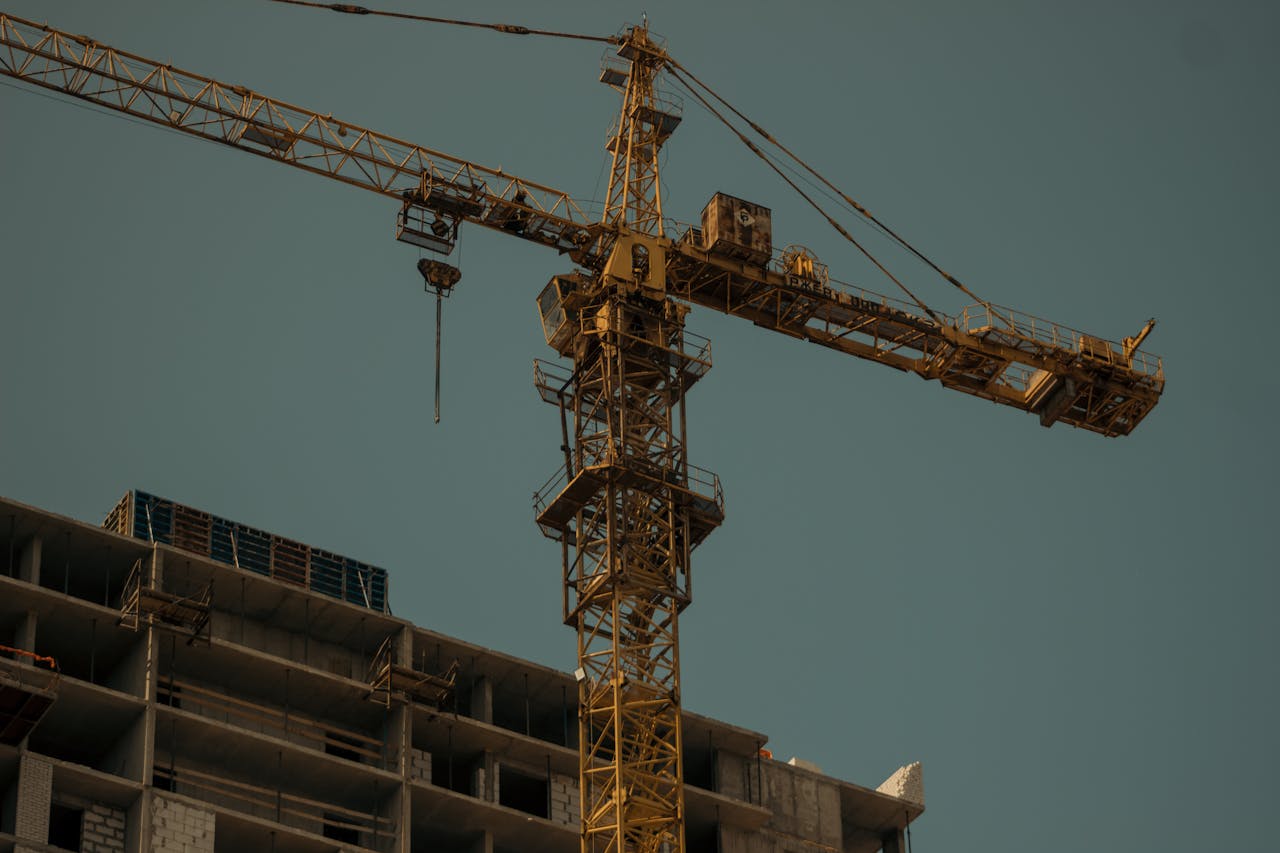 about-us-01 Vertical shot of a tower crane towering above an urban construction site, set against a plain sky.